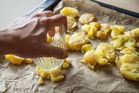 Christin Klose/dpa-tmn | Die gekochten Kartoffeln kommen auf ein Backblech und werden mit einem Glas zerdrückt. Mit Olivenöl bepinseln und Salzflocken bestreuen, dann 40 bis 45 Minuten knusprig backen.