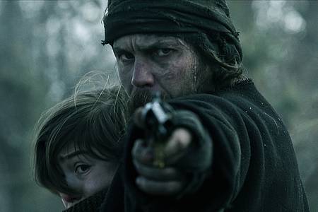 Ein bärtiger Mann drückt ein Kind an sich und richtet seinen Pistolenlauf auf die Kamera - American Primeval auf Netflix.