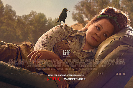 Melissa McCarthy liegt seitlich auf einem Feld, während ein Vogel auf ihrem Arm sitzt.
