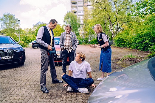 Drei Discounter-Mitarbeitende stehen auf einem Parkplatz in ihrer Mitte sitzt ein Kollege auf dem Boden