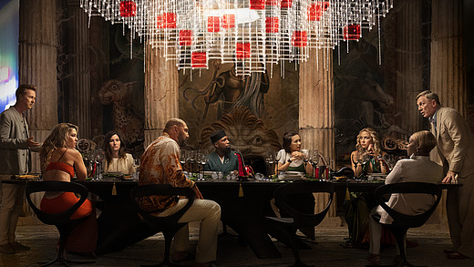 Alle Protagonisten stehen an einer langen Tafel in einem edlen Esszimmer.