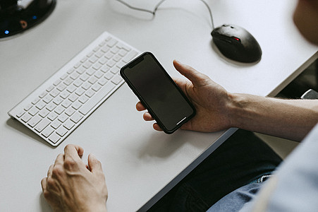 Hand hält schwarzes Smartphone, Schreibtisch und Tastatur im Hintergrund
