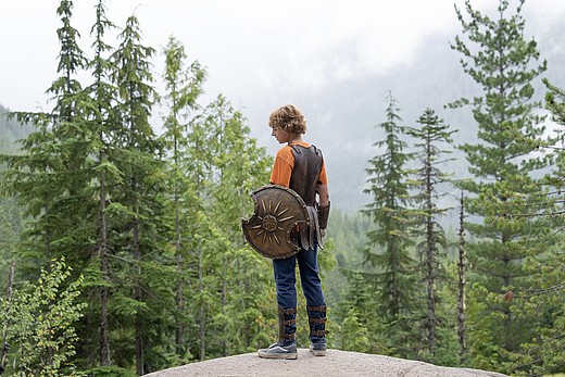 Percy Jackson aus der gleichnamigen Serie steht mit gezogenem Schwert auf einer Klippe im Wald.