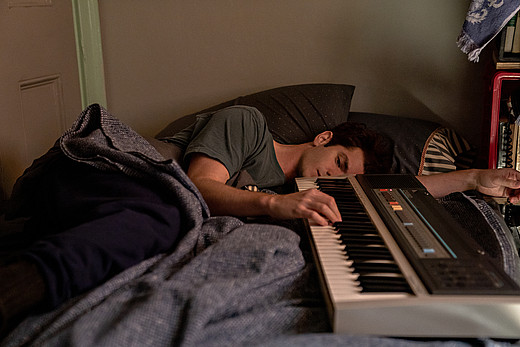 Andrew Garfield liegt mit einem Keyboard auf seinem Bett und drückt gedankenverloren die Tasten.