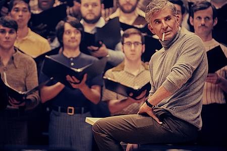 Filmszene: Leonard Bernstein in Maestro sitzt vor einem erwartungsvollen Chor. 